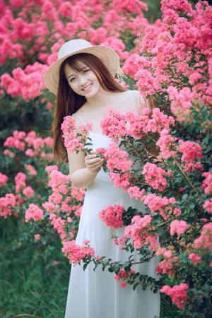 穿白色連衣裙和草帽的女人在粉紅色開花灌木叢中微笑。