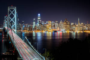 夜間景色，一座亮燈的橋樑在水面上留下燈光軌跡，面向發光的城市天際線。 — 天際線 壁紙
