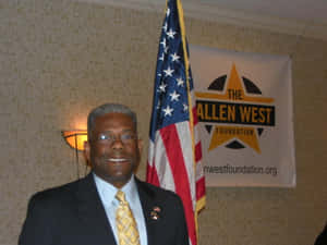 活動中穿著西裝嘅男士微笑，背後有美國國旗同Allen West Foundation橫額。