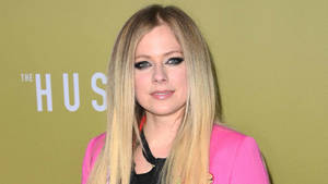 Avril Lavigne 在活動中穿著粉紅色西裝外套，背景為綠色。