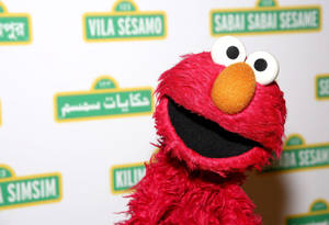 紅色毛絨絨的 Elmo 木偶微笑，背後有芝麻街標誌。