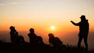 四個朋友在山頂上以日落為背景的剪影，一人指向地平線。 — 軍事動圖 壁紙