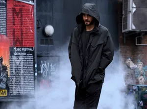 穿著黑色連帽雨衣的男人站在煙霧瀰漫的塗鴉小巷中,靠近音樂節海報。