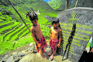 兩名身穿傳統服飾的年輕原住民男孩站在標誌附近，俯瞰梯田。