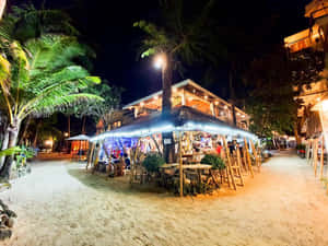 夜晚海灘酒吧，棕櫚樹和客人在燈串下享用飲品。 — 熱帶海灘 壁紙