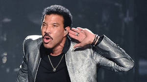 Lionel Richie 在舞台上唱歌，身穿銀色外套和黑色襯衫。