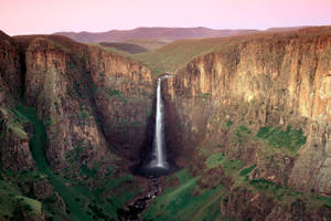 一道高瀑布在日落時分沿著陡峭的峽谷傾瀉而下，天空呈粉紅色，懸崖呈綠色。