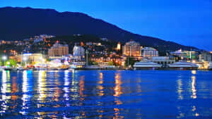 暮色中燈光在水面上閃爍的沿海城市，背景為山脈 — 城市景觀 壁紙