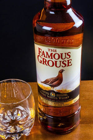 一瓶 Famous Grouse 威士忌和一個玻璃杯放在木桌上。