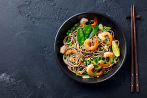 黑色碗中的蕎麥麵配蝦、荷蘭豆和青檸角，附筷子。