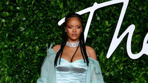 Rihanna 身穿藍色緞面長袍，辮子和頸鍊，背景是綠色樹籬。