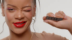 Rihanna 眨眼，手持Fenty唇膏，搭配大膽紅唇和粉紅色眼線。