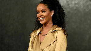 Rihanna 喺深色背景前著住金色 trench coat 微笑。
