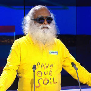 白鬍子戴太陽眼鏡的男人穿著黃色「Save Our Soil」襯衫站在講台。