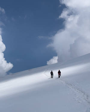 兩人在多雲的天空下滑雪上山。 — 冬天桌面 壁紙