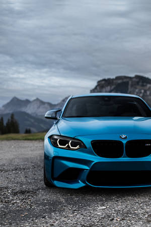 多雲天空下，藍色BMW M2跑車停泊在碎石上靠近山脈 — 寶馬 iPhone 壁紙