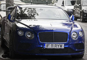 一輛藍色 Bentley Continental GT 停泊在濕漉漉的城市街道上，帶有樹木倒影。