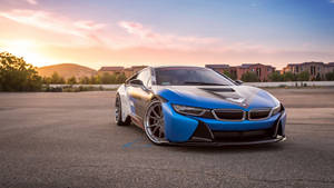 藍色BMW i8跑車停在夕陽下的瀝青路上，背景有房屋。 — 日出 壁紙