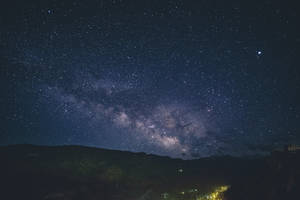 銀河系在黑暗山脈和燈火通明的村莊上方的星空夜空中延伸。 — 星空夜 壁紙