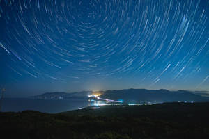 星軌在夜間沿海小鎮上空劃出弧線，遠處燈光沿著海岸線。 — 銀河系 壁紙