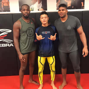 三位男性格鬥選手喺健身室入面，喺紅色軟墊同海報前擺甫士。