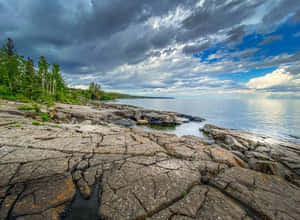 裂縫的岩石海岸在戲劇性的多雲天空下與平靜的湖泊相遇，有綠樹。 — 湖 壁紙
