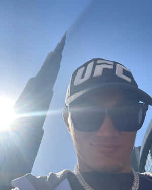 戴住UFC帽同太陽眼鏡嘅人喺哈利法塔附近微笑，有太陽眩光。