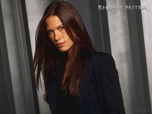 Rhona Mitra 身穿深色西裝外套，背景為灰色垂直條紋。
