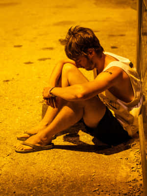 一個年輕男子在夜晚獨自坐在人行道上，在黃色街燈下低頭。