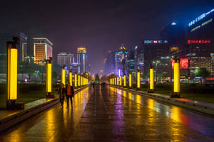 夜晚城市步道，發光的黃色柱子和摩天大樓在潮濕的路面上反射。 — 唐人街 GIF 壁紙