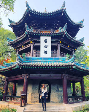女士站在裝飾華麗的中式亭閣入口，有彎曲屋頂和綠植。 — 唐人街 GIF 壁紙