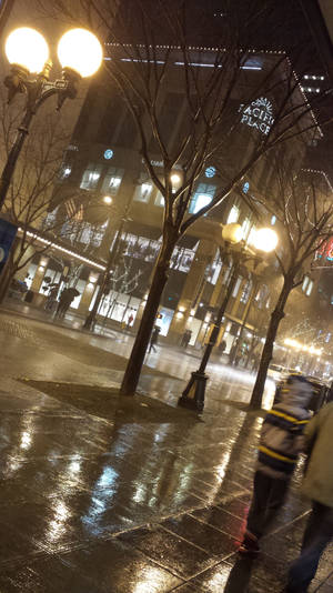 行人夜晚在發光的街燈下走在被雨水浸濕的城市人行道上。