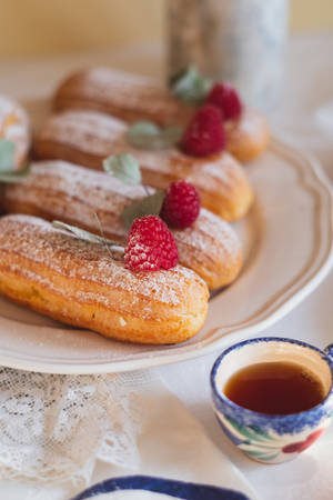 一排糖粉泡芙配上覆盆子和鼠尾草葉，旁邊有一杯花卉圖案的茶。