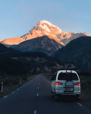白色貨車在山路上接近夕陽下的雪山頂峰。 — 冬天桌面 壁紙