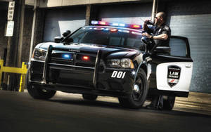 身穿黑色制服的警員從黑色道奇警車的打開車門探出身來，警車在小巷中閃著警燈。
