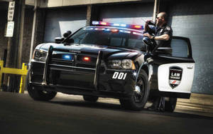 身穿黑色制服的警員從黑色道奇警車打開的車門探出身子，警車在小巷中閃著警燈。