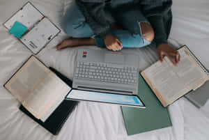 一個人在床上用筆記型電腦和周圍打開的書本學習。