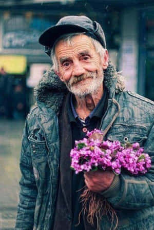 一位穿著冬季外套的長者在城市街道上微笑著手持紫色花朵。 — 美學心形 壁紙