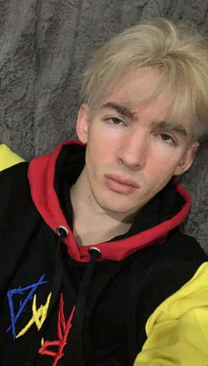 年輕金髮男子穿著彩色連帽衫，背景為灰色牆壁。