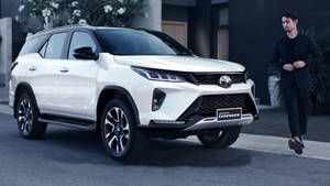 一架白色豐田 Fortuner Legend SUV 停泊在現代街道上，附近有一位身穿黑色西裝的男子走過。