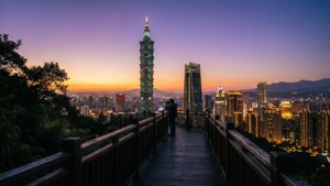 人站在木製觀景台上拍攝日落時分的台北城市景觀，台北101亮起燈光