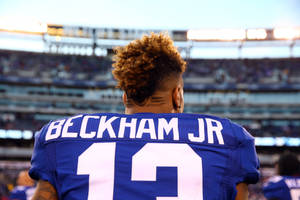 美式足球員 Beckham Jr 身穿藍色球衣站在夕陽下的球場。 — 奧德爾·貝克漢姆 Jr 壁紙