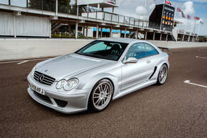 銀色 Mercedes-Benz CLK GTR 賽車在賽道上，背景有看台和旗幟。