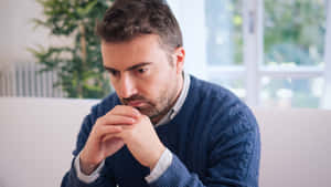 身穿藍色毛衣的男子坐在室內，雙手交疊靠近下巴，顯得若有所思。