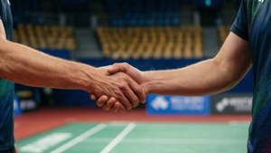 兩名羽毛球運動員在場上握手，身後是空的黃色座位。