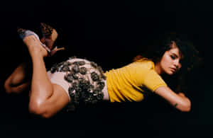 一位身穿黃色上衣和亮片裙的女性，在黑色背景下穿著圖案高跟鞋擺姿勢。