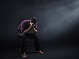 一個男人坐在凳子上，在黑暗的房間裡用聚光燈照射下掩面。