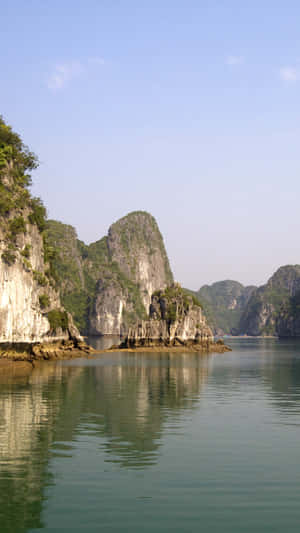 寧靜的海灣，高聳的石灰岩喀斯特地貌倒映在平靜的碧綠海水中，天空晴朗湛藍。
