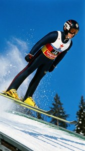 冬季比賽中，滑雪跳躍選手在藍天和積雪斜坡的背景下半空中。
