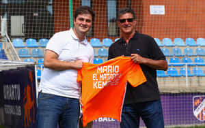 兩名男子在體育場舉起一件印有「Mario El Matador Kempes」的橙色足球球衣。